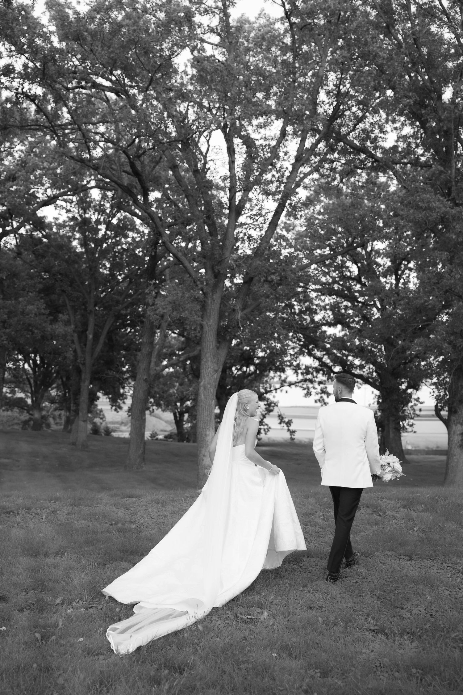 Black and white photo of a bride and groom walking hand in hand through a tree-lined grassy area.