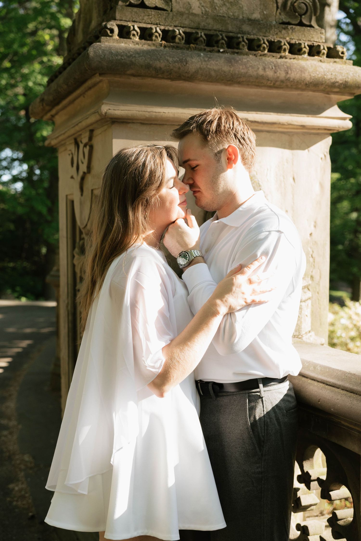 Couple in white attire embrace closely beside ornate stone column in sunny park.