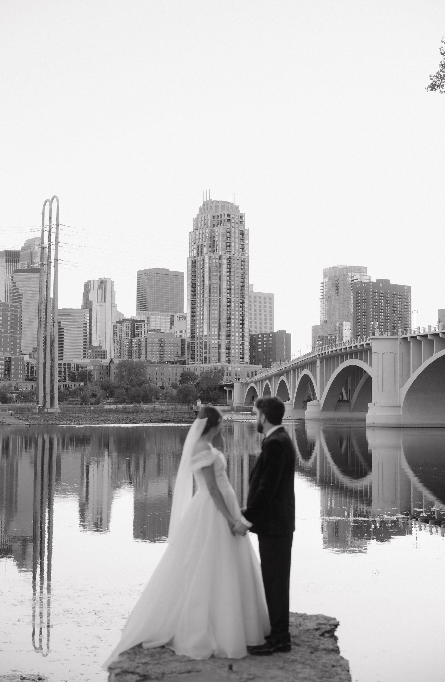 Black and white wedding portrait of a couple standing by a river with a city skyline and bridge.