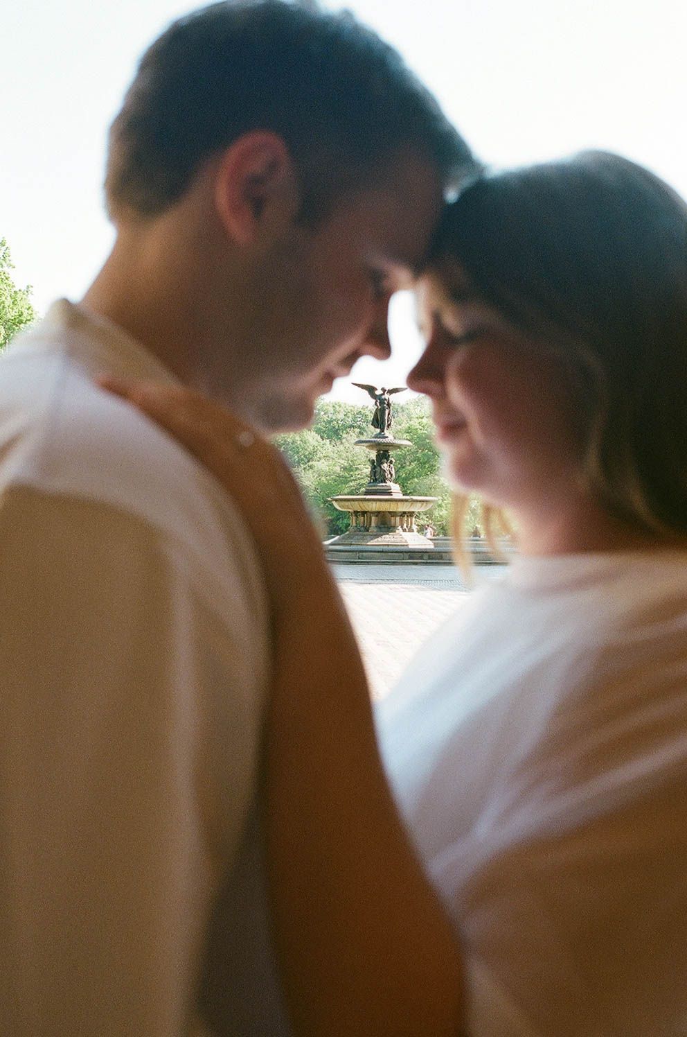 Romantic close-up of couple touching foreheads with Bethesda Fountain softly blurred in background.