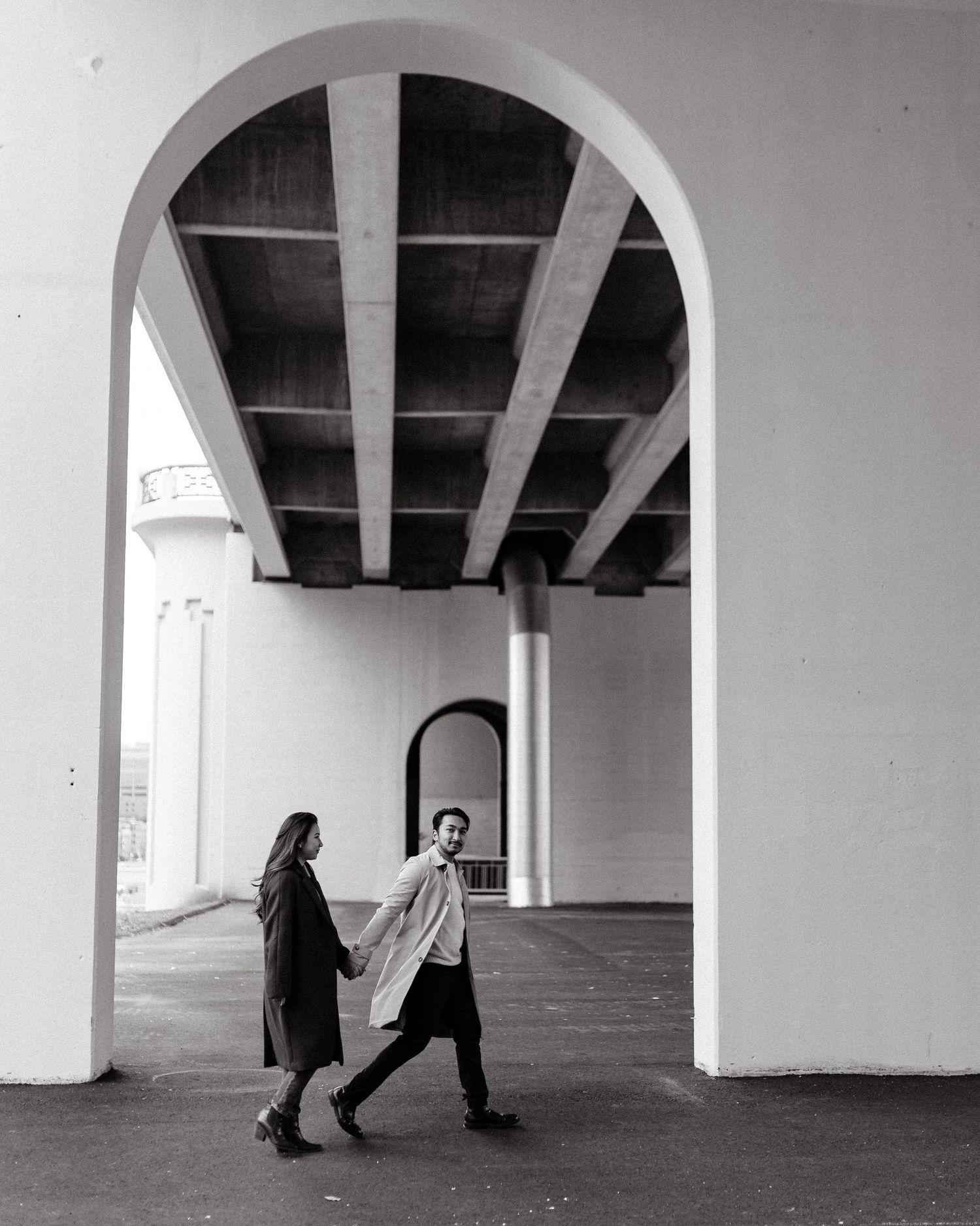 Black and white photo of a couple holding hands and walking through a grand white arched walkway.