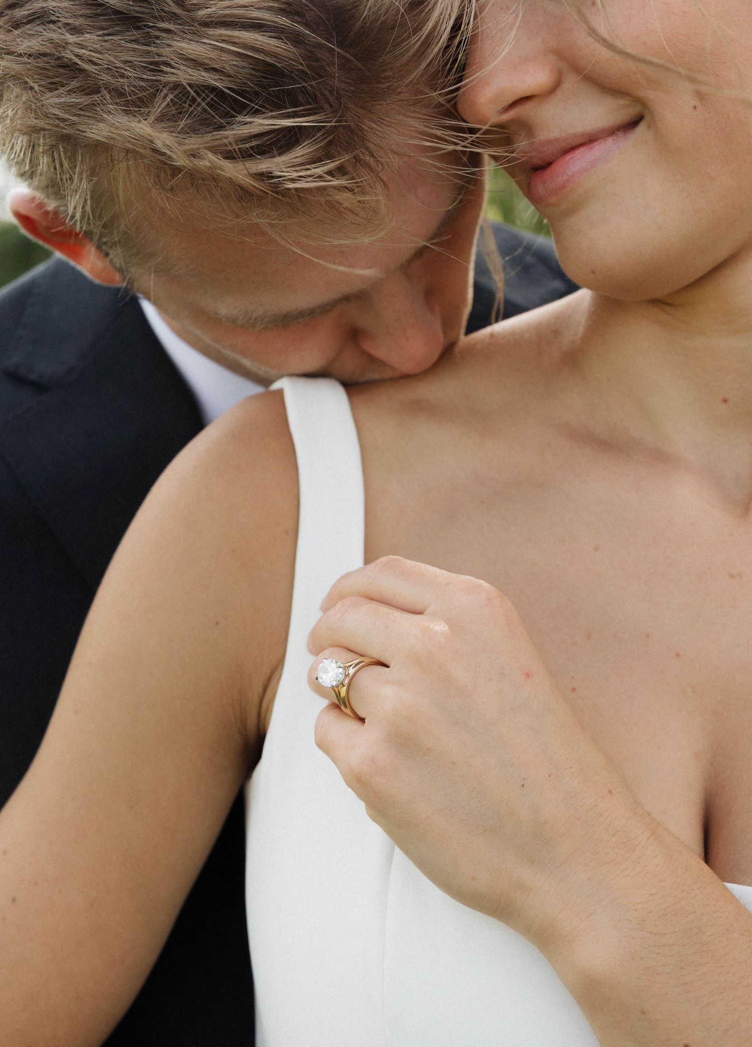 Groom kisses bride's shoulder as she smiles showing diamond engagement and wedding rings.