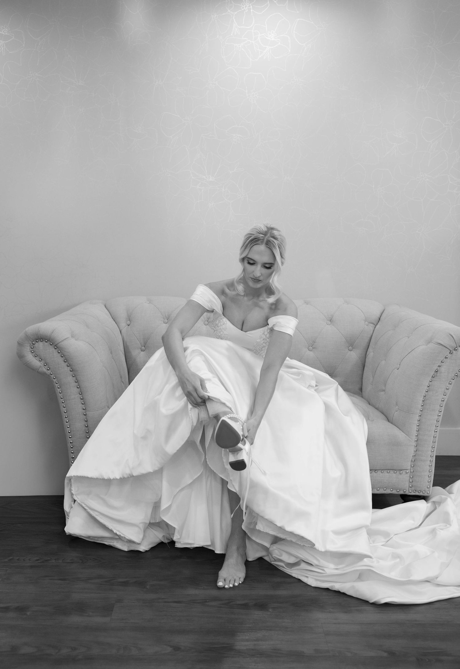 Black and white photo of a bride in an off-shoulder gown adjusting her shoe while seated on a tufted sofa.