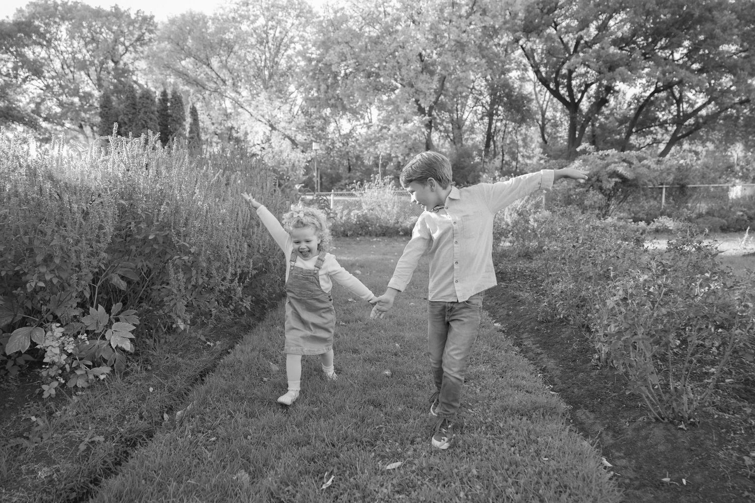 Two young children hold hands dancing along garden path surrounded by lush green plants in black and white.