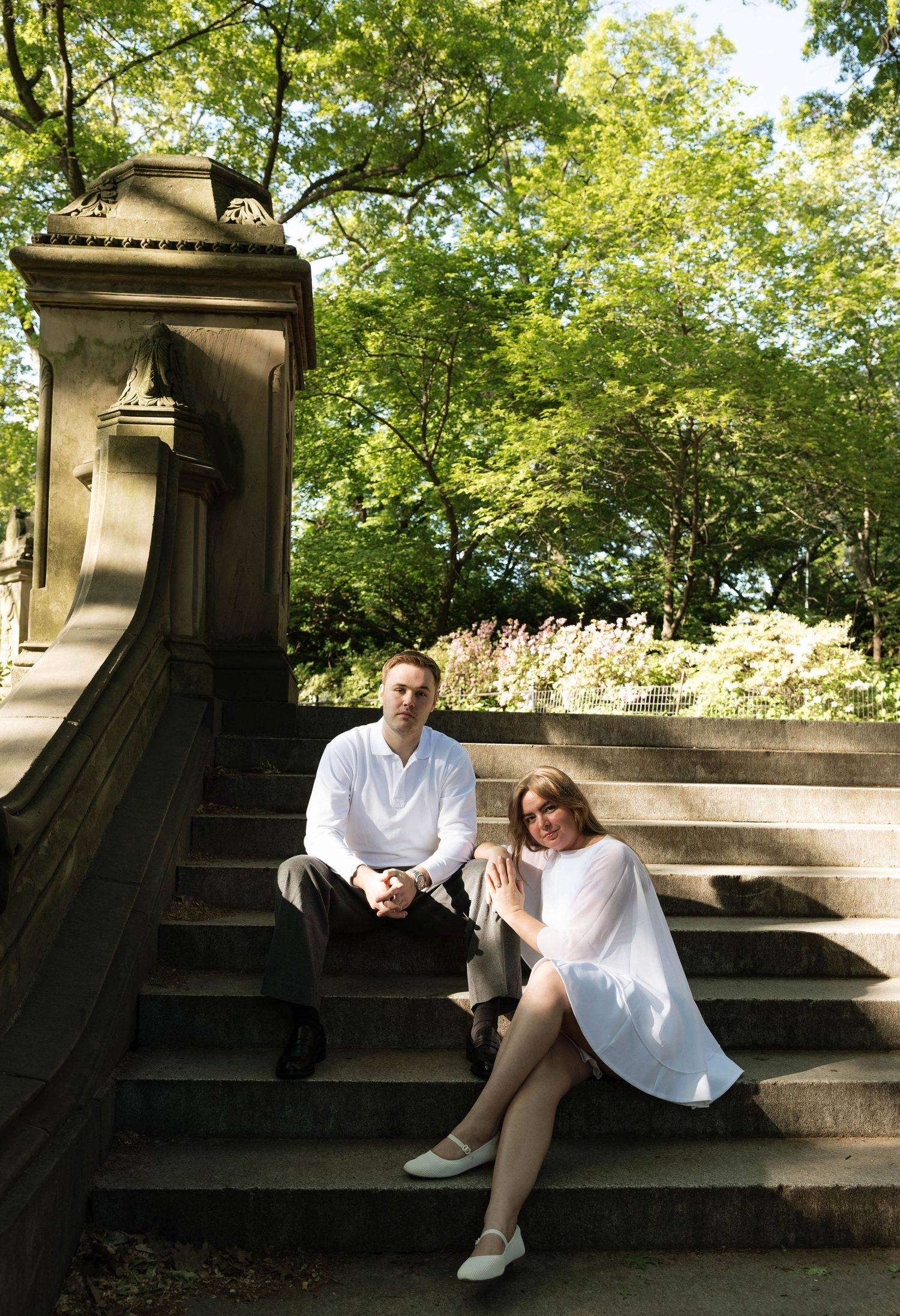 Couple in white outfits sitting on stone steps surrounded by lush green trees in Central Park.