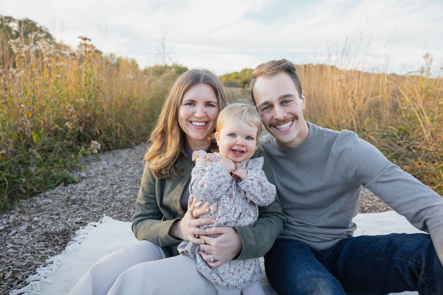 Happy family with baby sitting outdoors on a gravel path surrounded by tall grass.