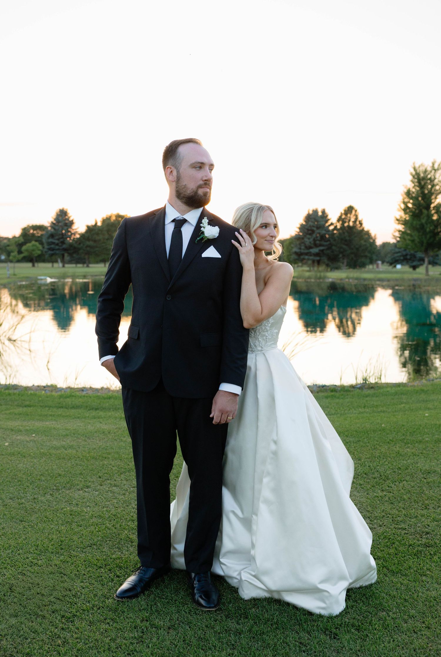 Bride and groom posing elegantly by a serene pond at golden hour during outdoor wedding.