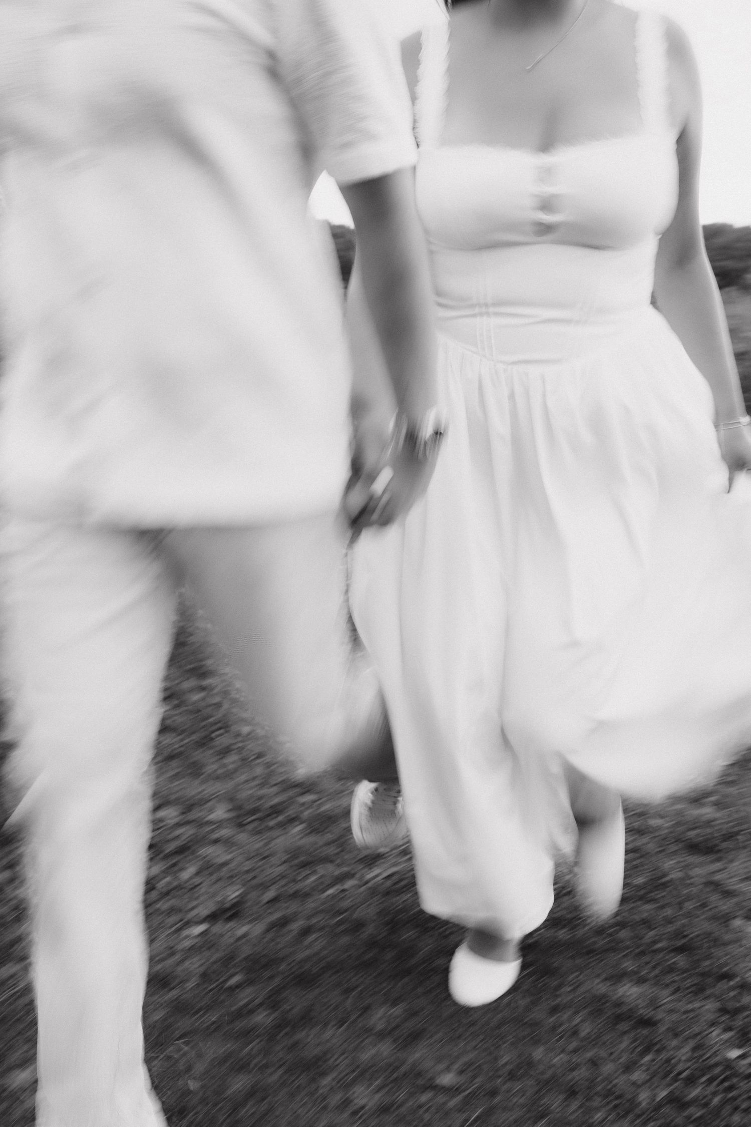 Motion blur black and white image of a couple in white clothing running hand in hand across a field.