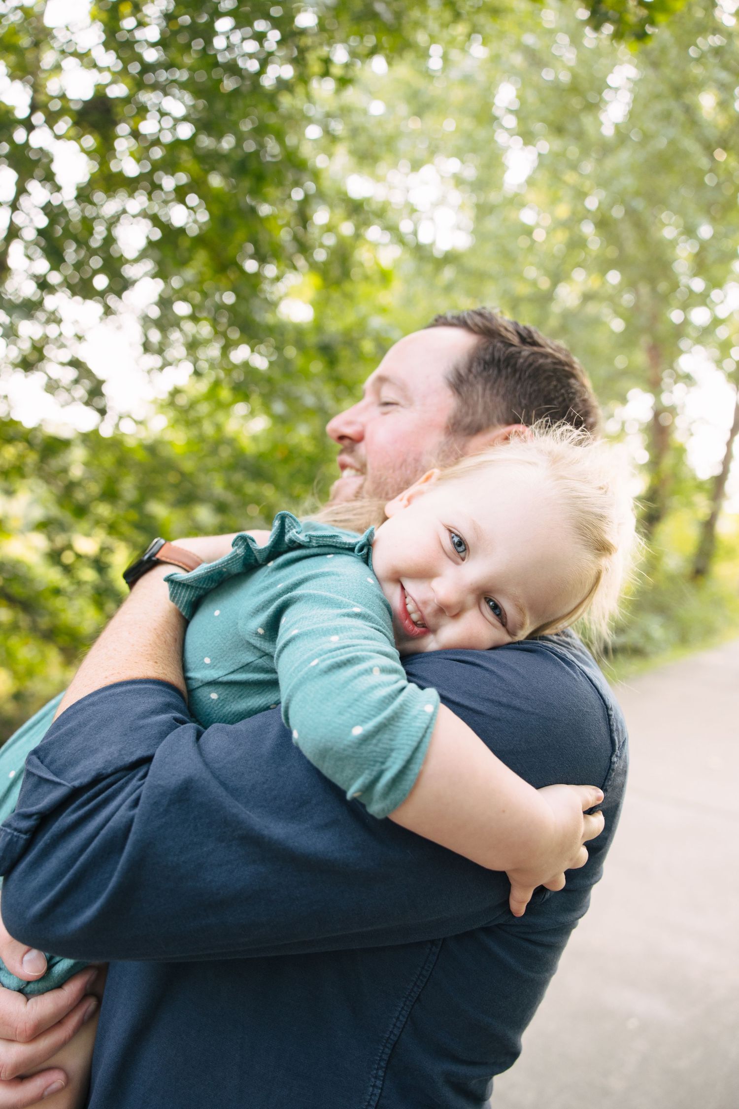 A bearded father tenderly hugs his smiling blonde toddler outdoors among lush green trees.
