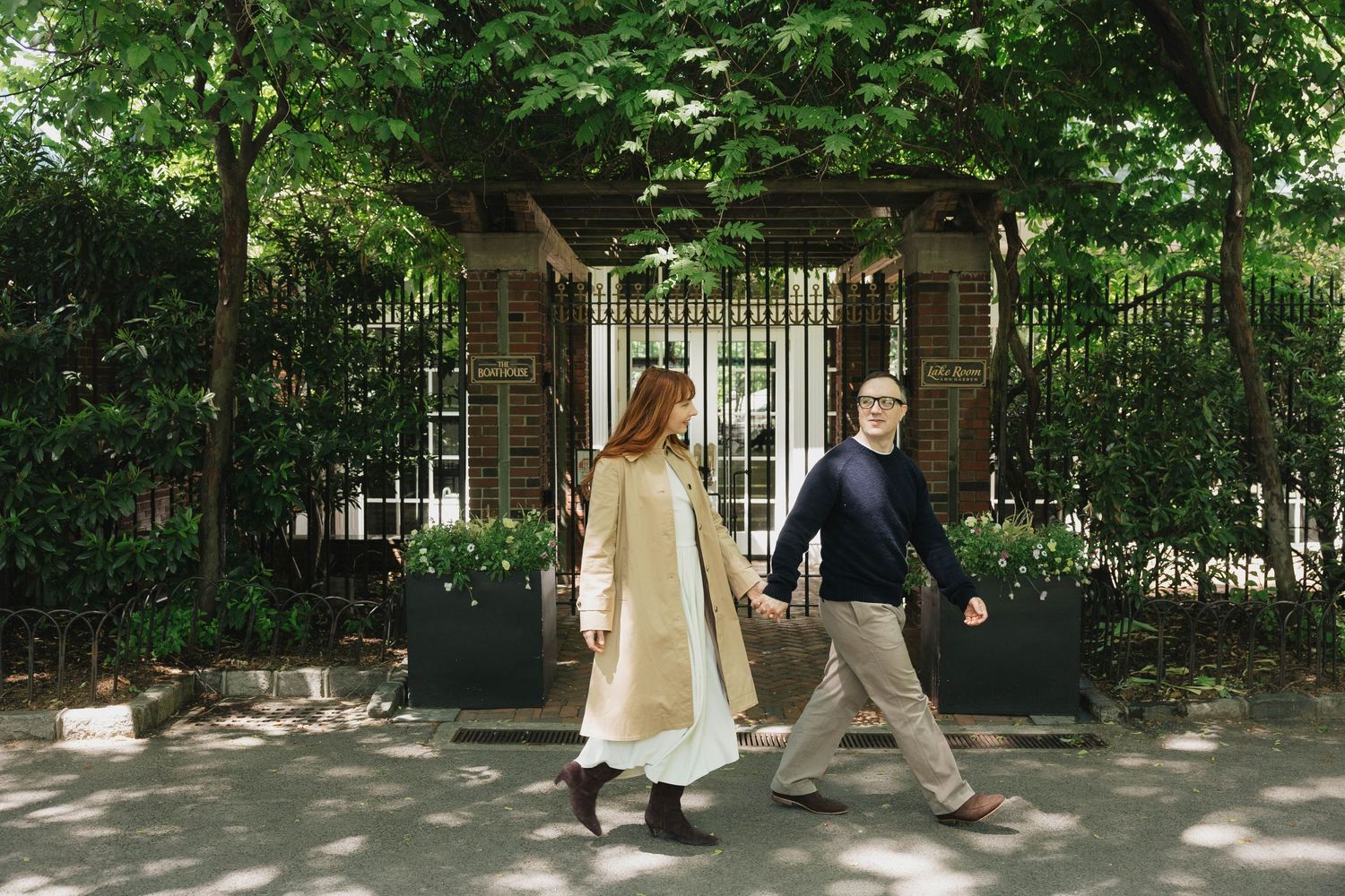 Couple walking hand in hand past a vine-covered pergola and iron gate in a sunny urban park setting.