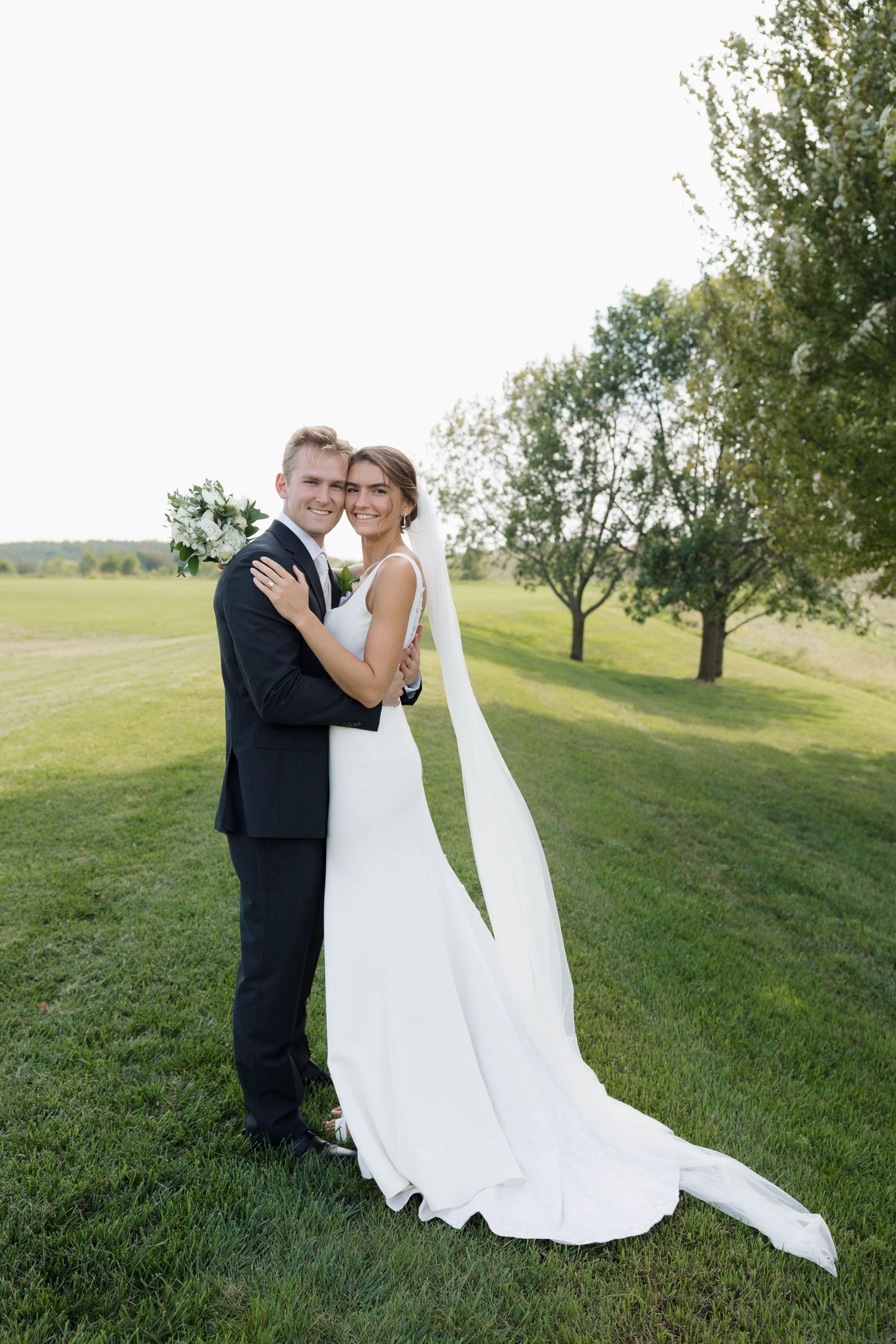 A bride and groom embrace on a green lawn, she in a flowing white gown with veil, he in a black tuxedo.