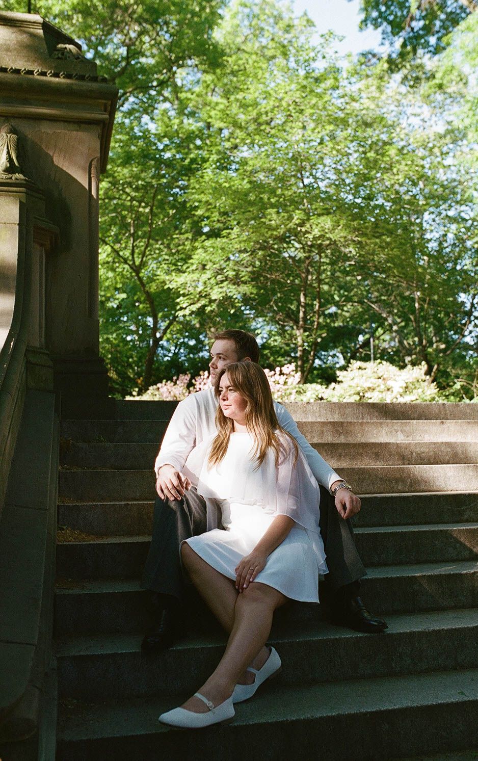 Couple sitting together on sunlit stone steps surrounded by lush green trees in Central Park.