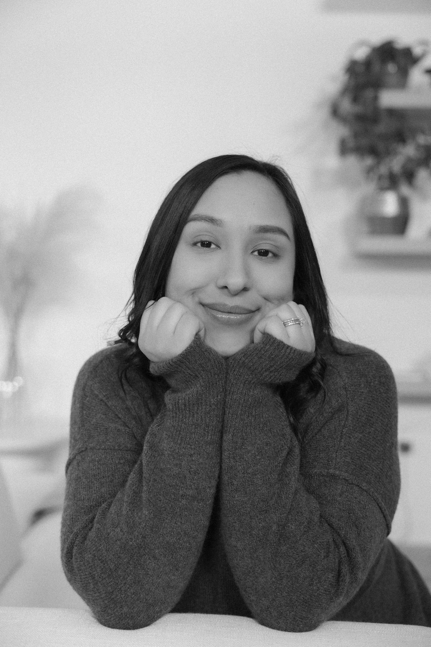 Black and white portrait of a smiling young woman in a cozy sweater resting her chin on her hands.