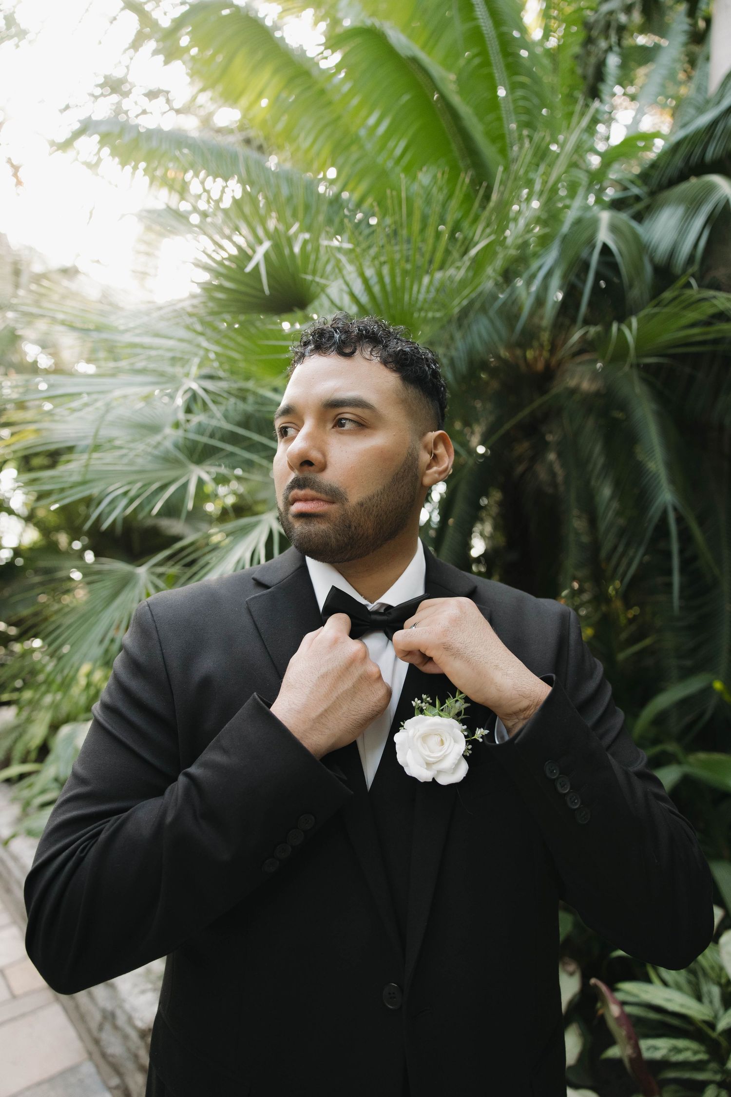 Groom in black tuxedo adjusts bow tie with white boutonniere among tropical plants.