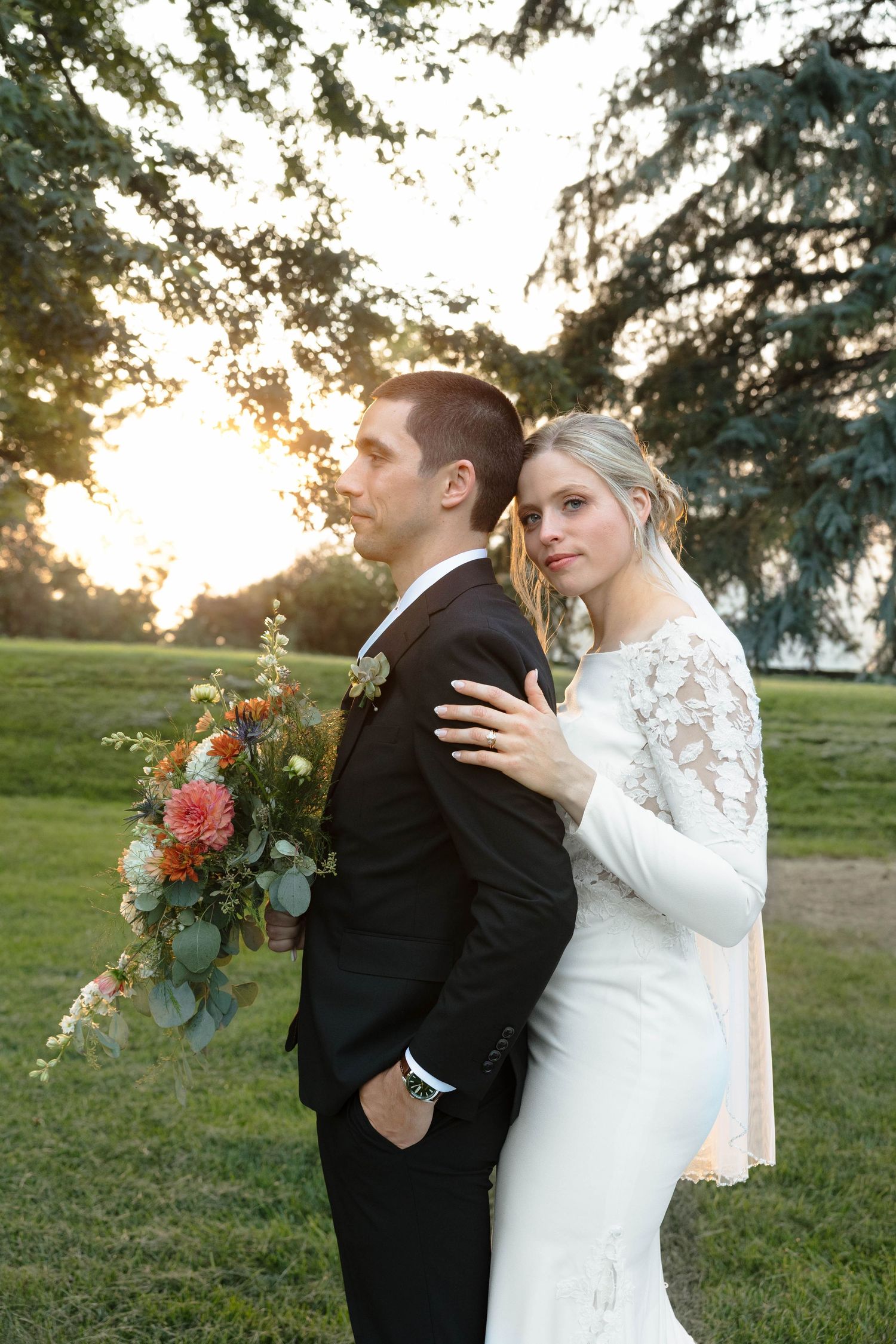 Bride in lace gown embraces groom holding bouquet at golden hour sunset outdoors.