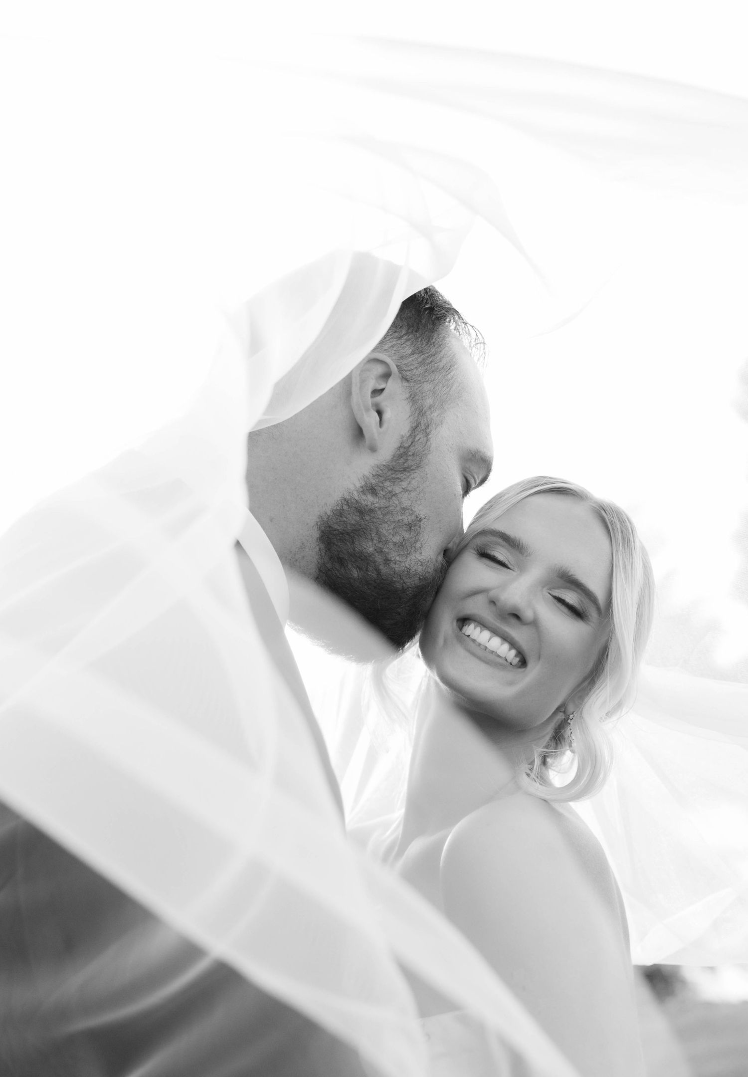 Black and white photo of a joyful bride laughing as the groom kisses her cheek beneath a flowing wedding veil.