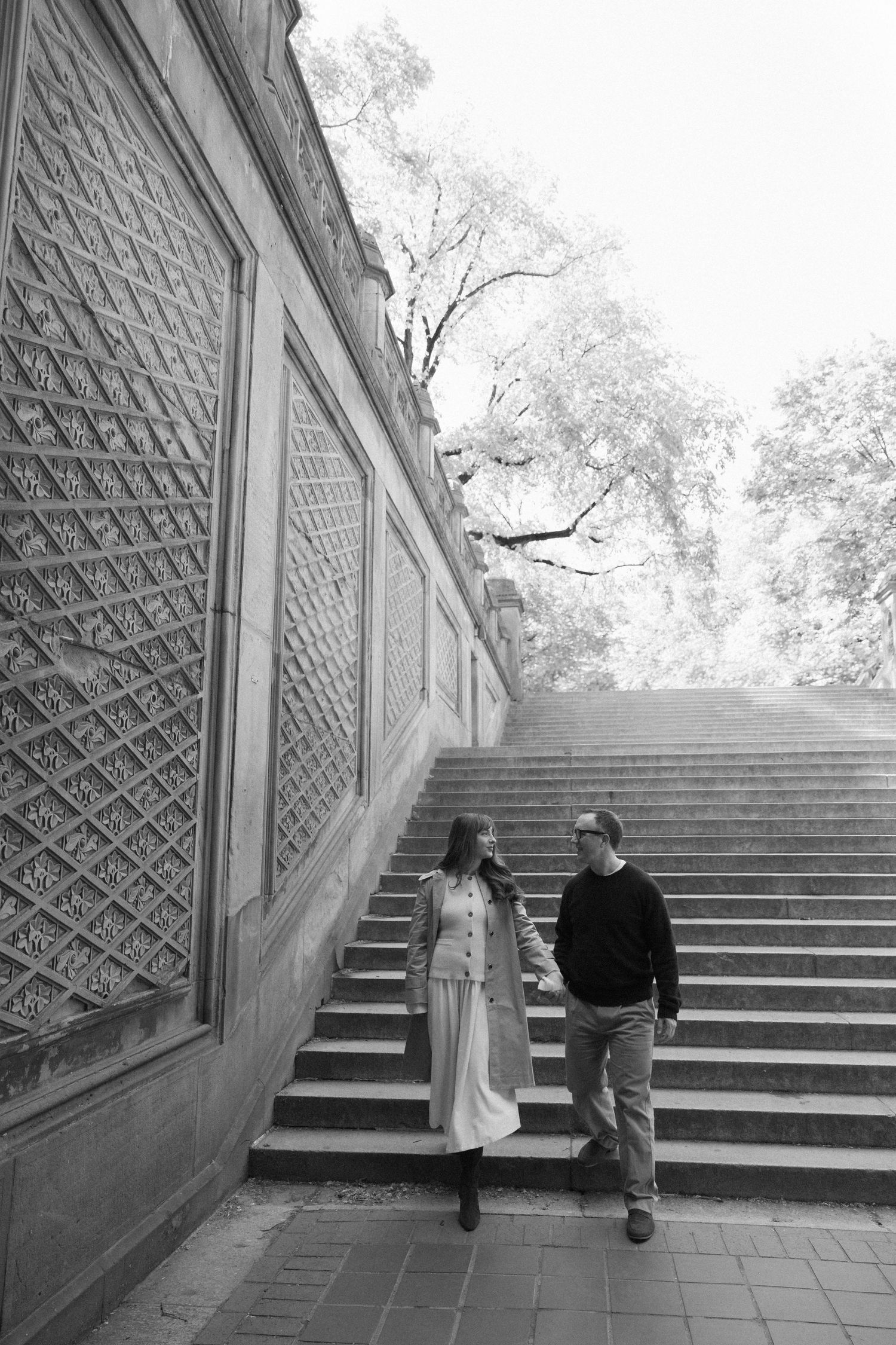 Black and white image of couple holding hands walking beside ornate stone wall and staircase in Central Park.