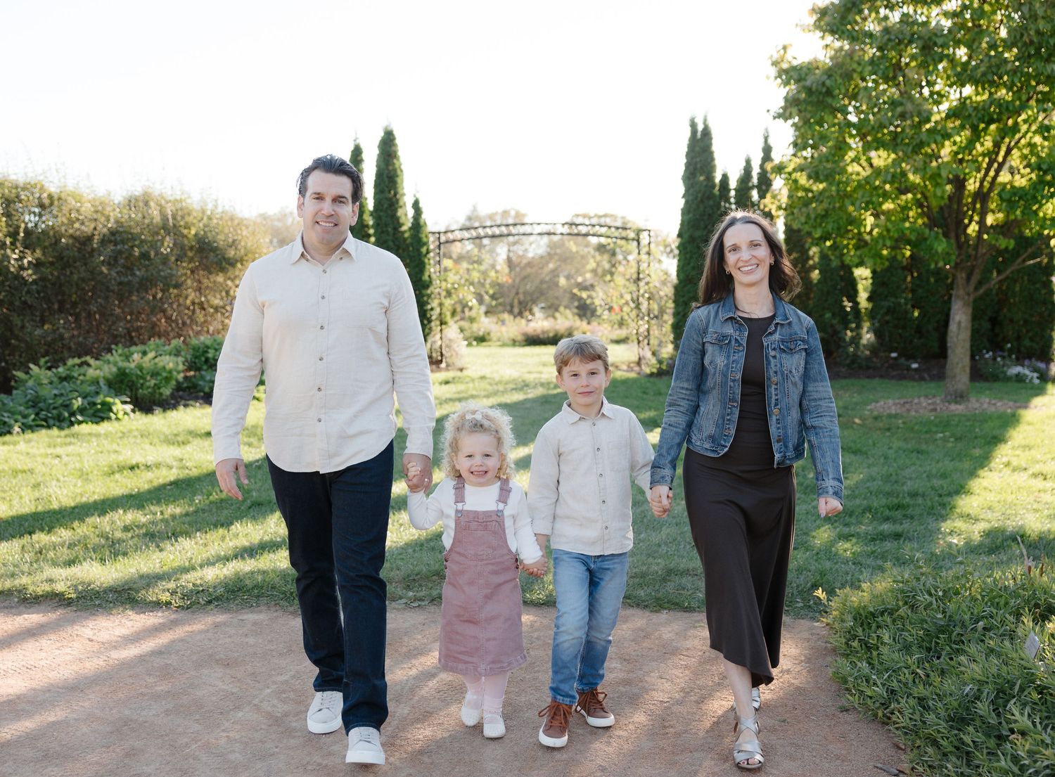 Happy family of four holding hands walking on garden path surrounded by green trees and landscaping.