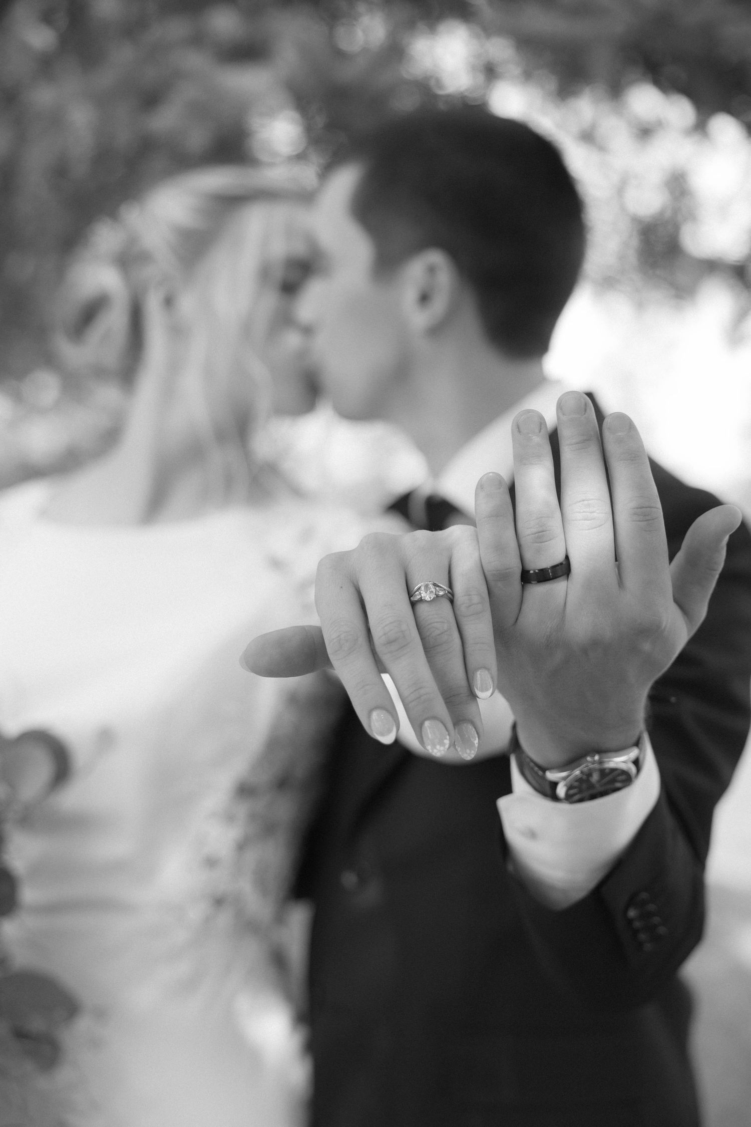 Black and white close-up of newlyweds showing off wedding rings while kissing softly in a blurred outdoor background.