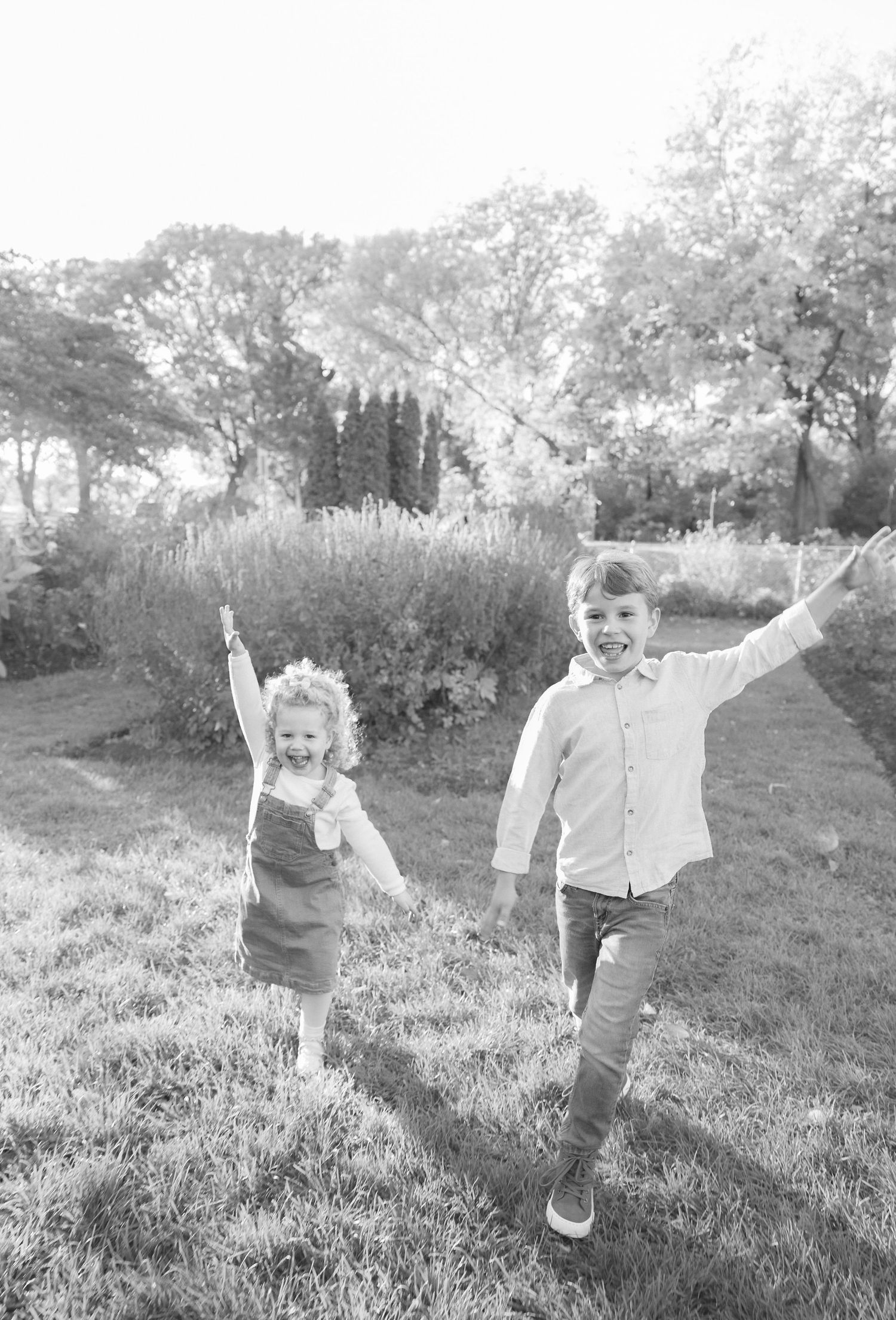Two joyful children run with arms raised in a black and white garden photo.