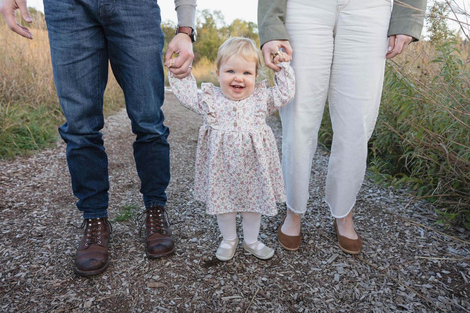 Baby girl in floral dress holding parents' hands while standing on a gravel path.