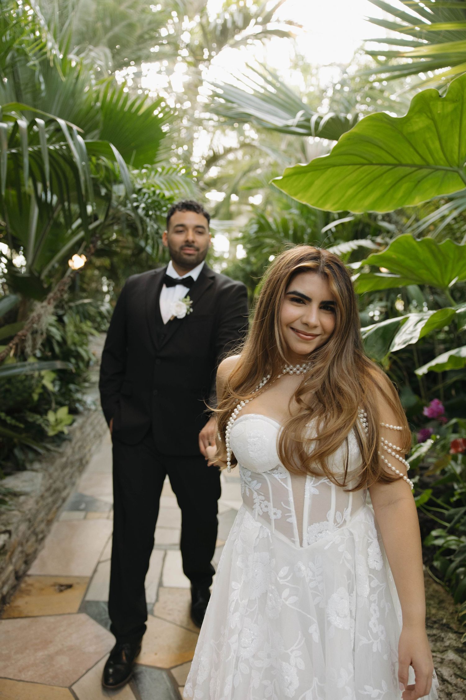 Bride in pearl-adorned gown leads groom in black tuxedo through lush tropical setting.
