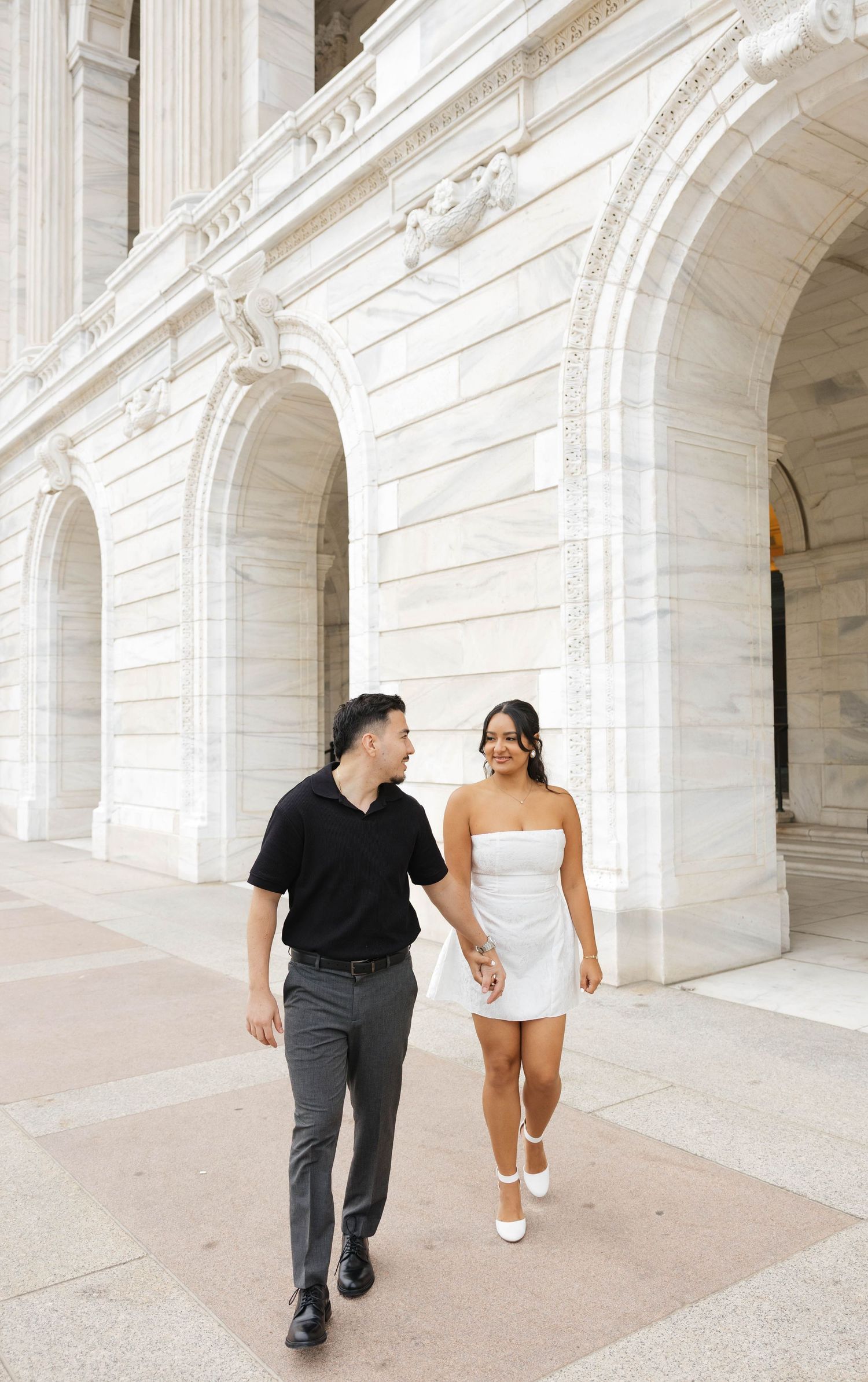 Smiling couple walking hand in hand past grand white marble arched columns of a neoclassical building.