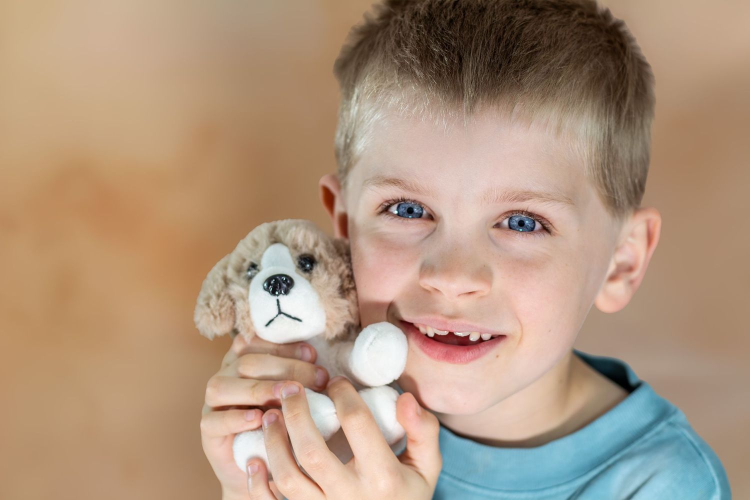 Smiling boy with blue eyes holding a small stuffed puppy toy, showing a missing tooth, against a warm beige background.