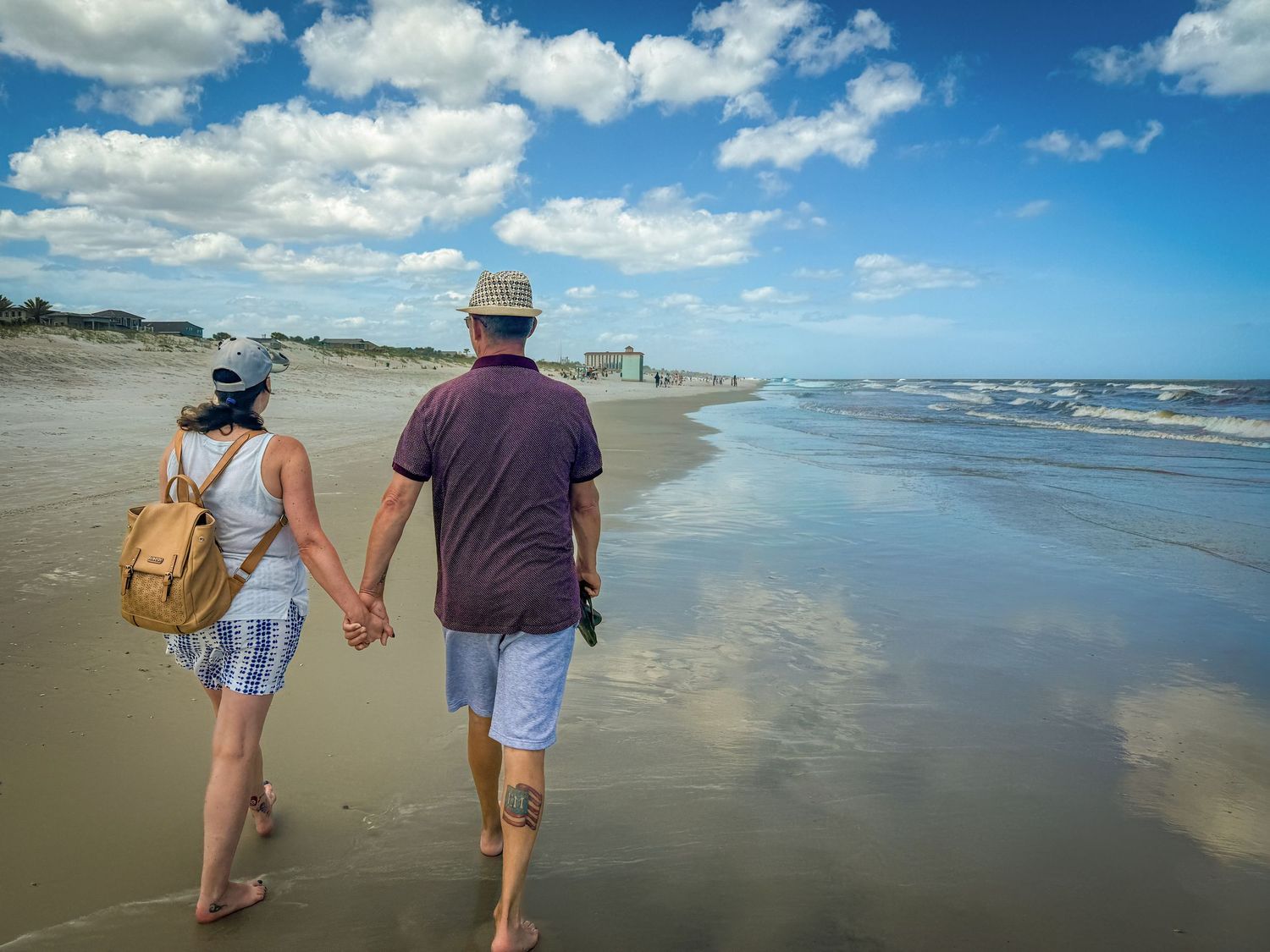 People walking along a wide sandy beach under blue skies with white puffy clouds.