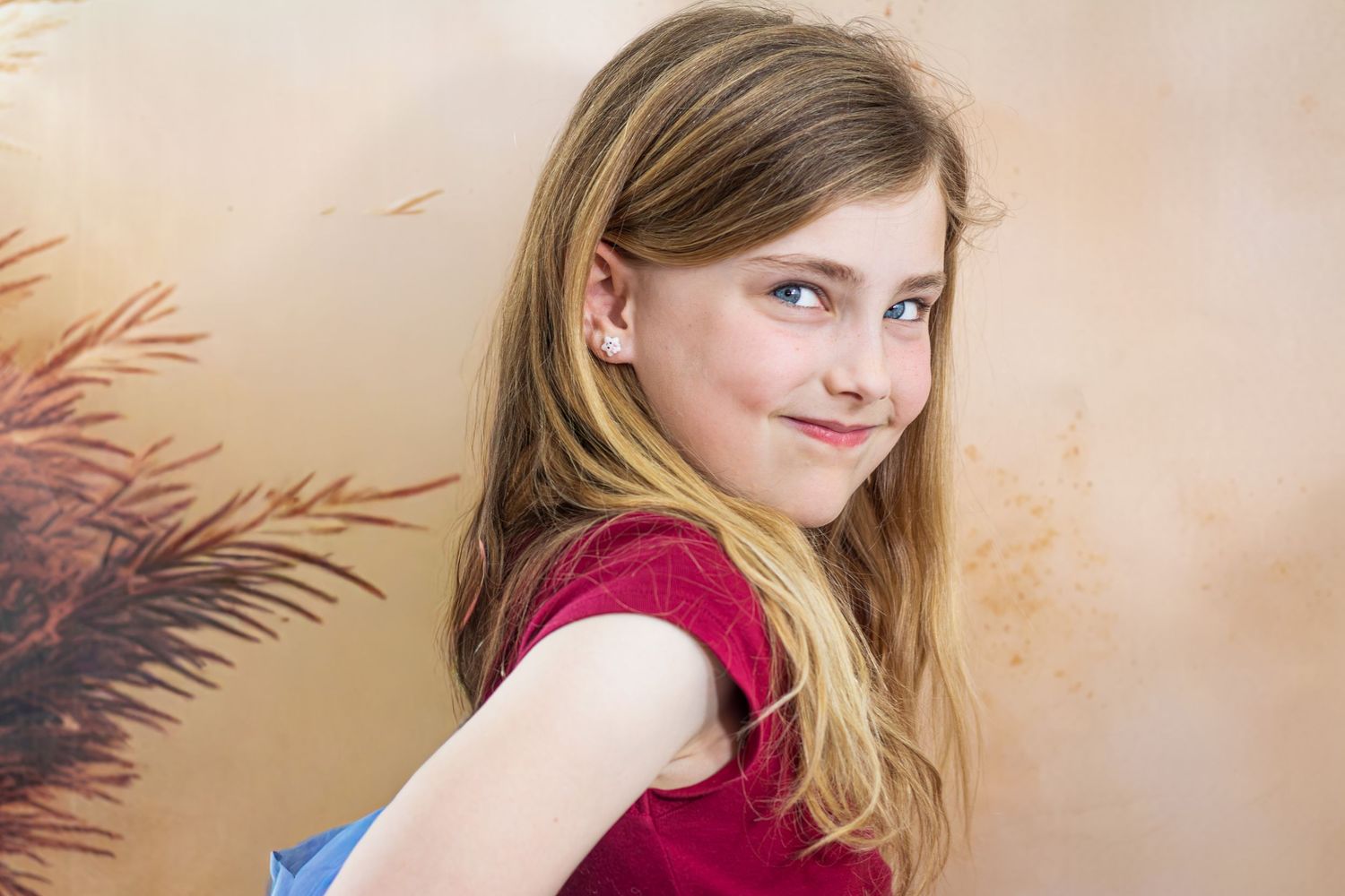 Young girl with long blonde hair and blue eyes smiling over shoulder in red top against beige background.