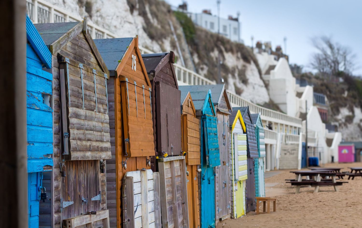 Row of colorful beach huts along a sandy beach with white cliffs in the background.