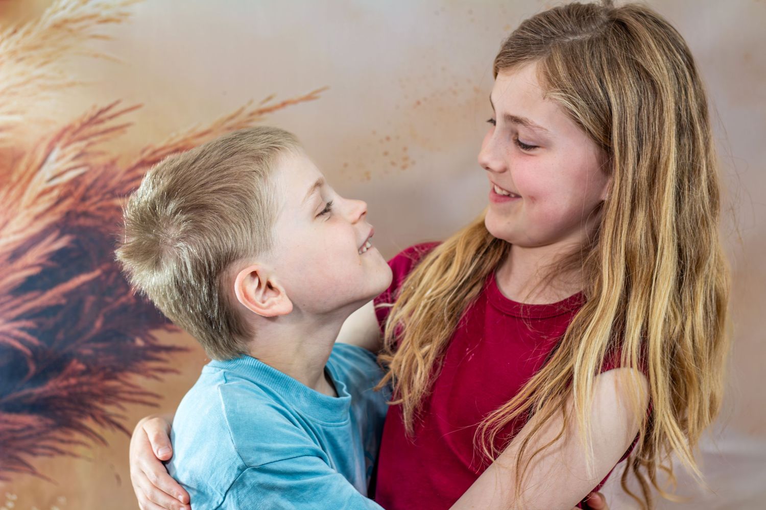 Young girl in red top hugging a small blonde boy in blue shirt, both smiling at each other.