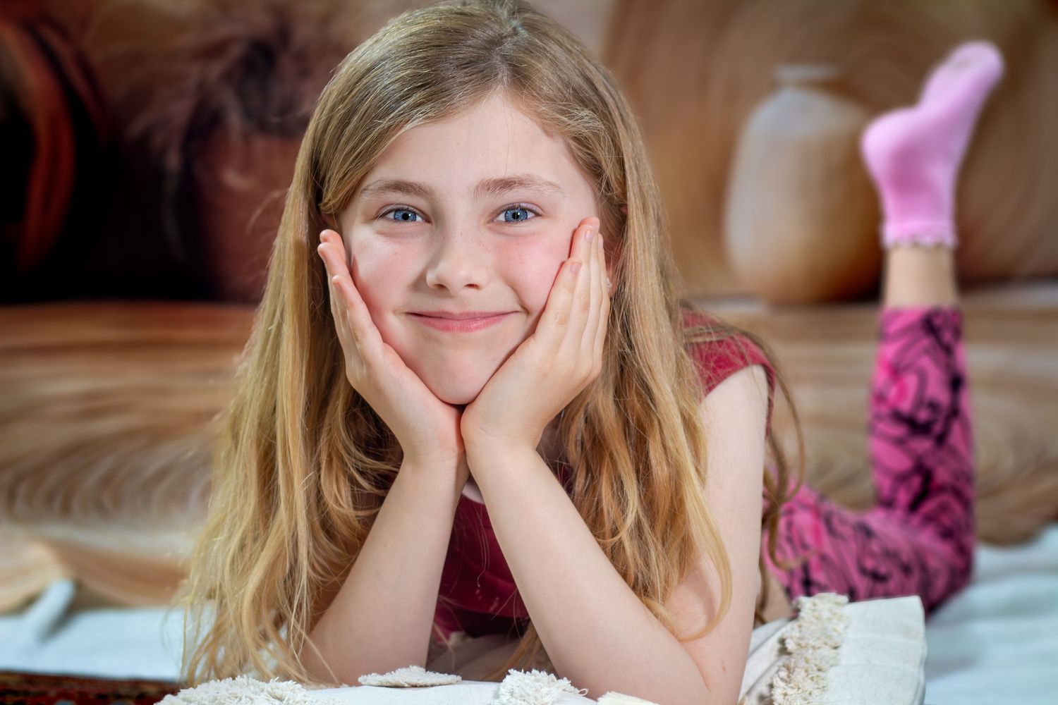 Young girl with long blonde hair resting her chin on hands, smiling while lying on floor near a brown leather sofa.