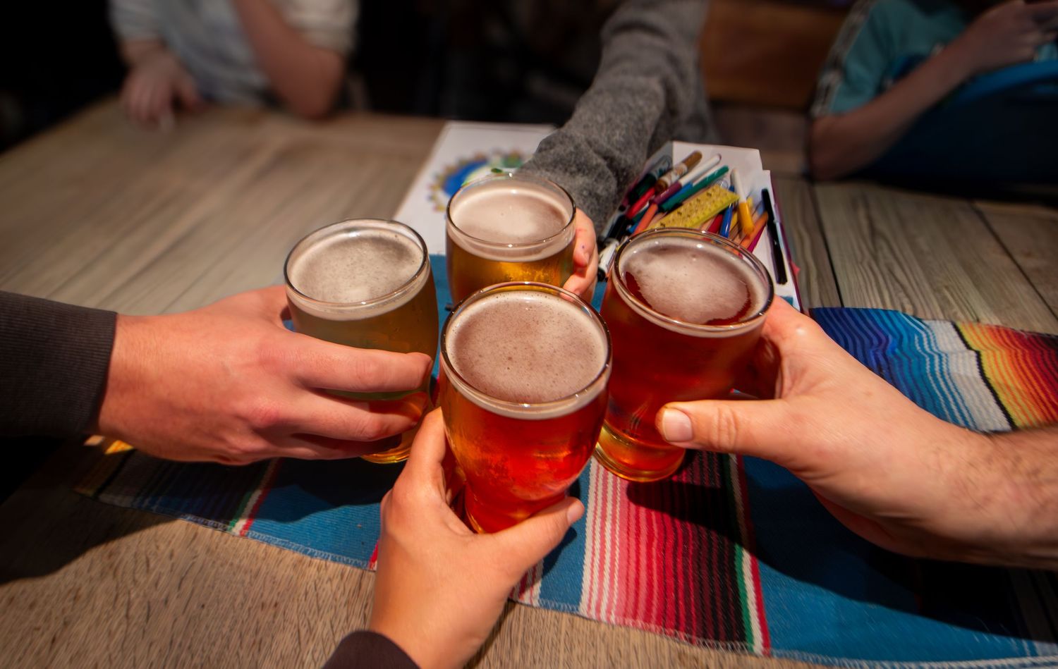 Hands toasting with beer glasses over a colorful striped table runner.