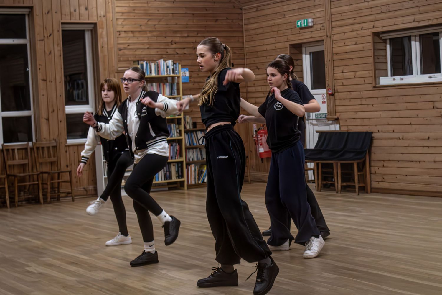 Four young women rehearse a dynamic choreographed dance routine in a wooden community hall.