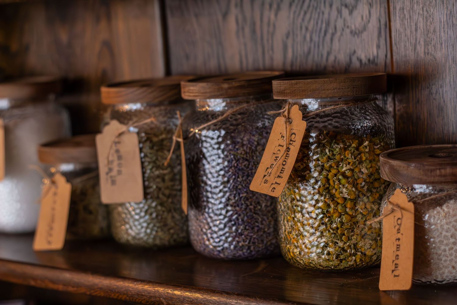 Rustic glass jars with handwritten labels displayed on a wooden shelf in a dimly lit setting.