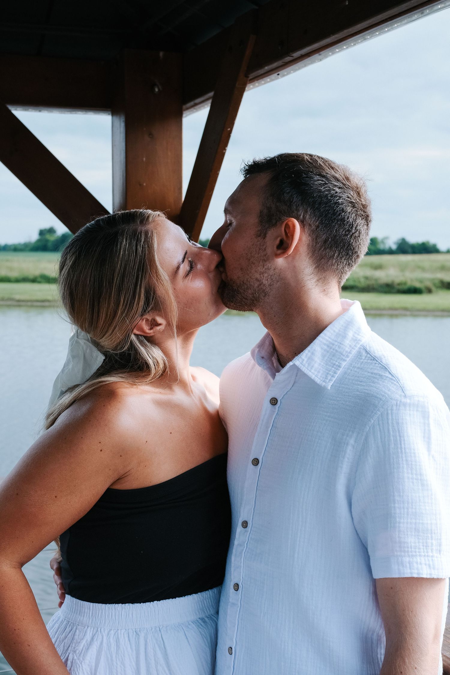 A couple shares a romantic kiss on a waterfront dock under a wooden pavilion structure.