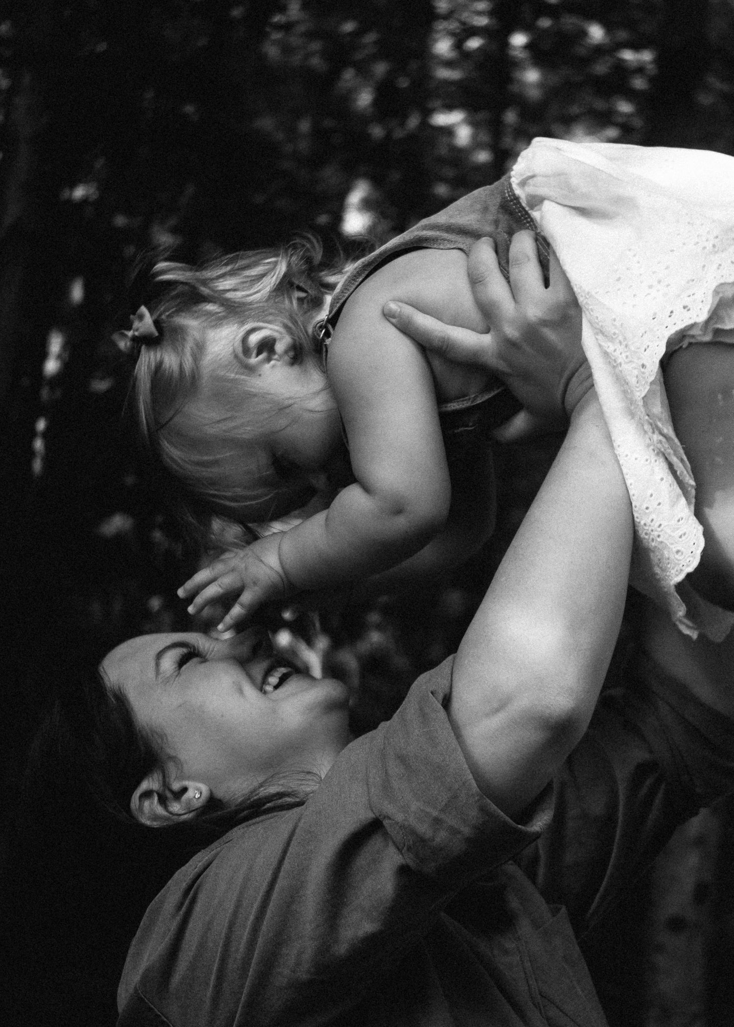 Black and white photo of a joyful mother lifting her laughing toddler daughter high in the air outdoors.