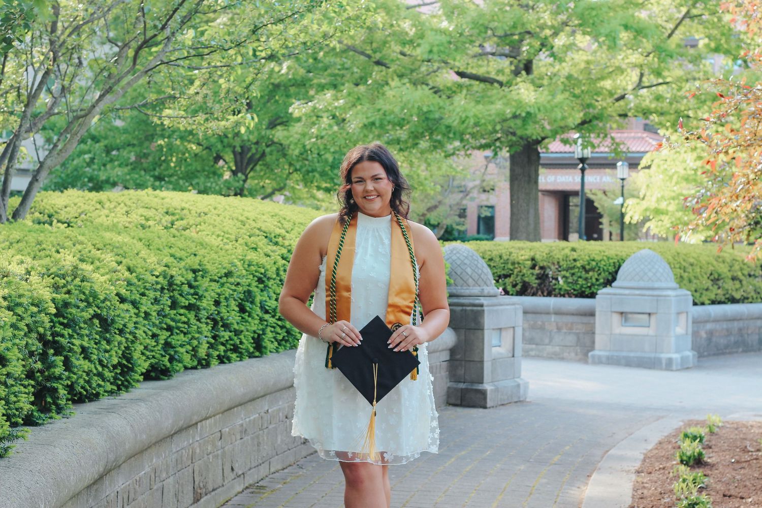 Smiling brunette woman in white dress with gold graduation stole holding cap outdoors on Purdue's college campus in West Lafayette