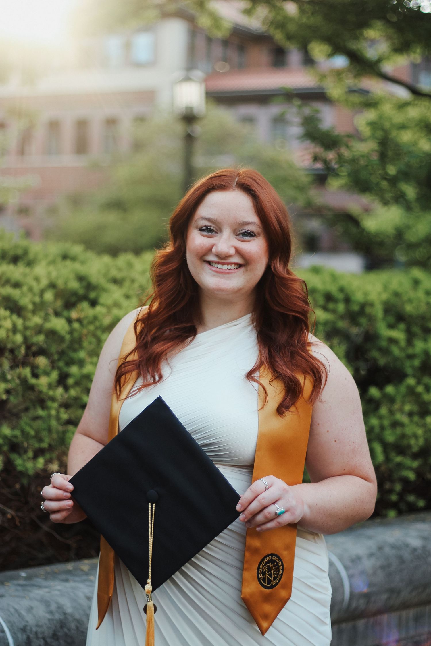 Smiling redhead woman in white dress holding graduation cap with gold stole on campus on Purdue's college campus in West Lafayette