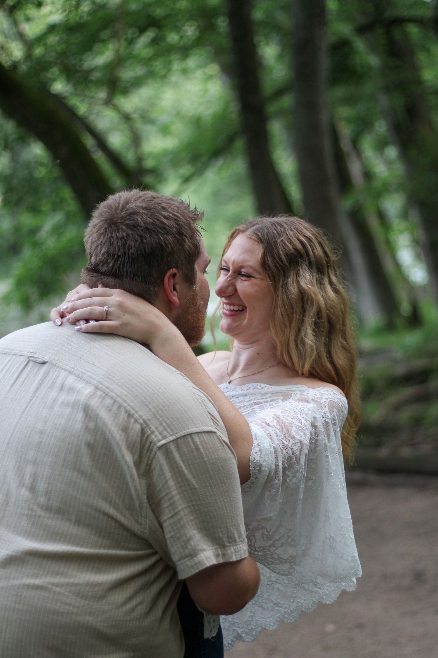 A smiling woman in a white lace top embraces a man in a beige shirt in a lush green forest setting.
