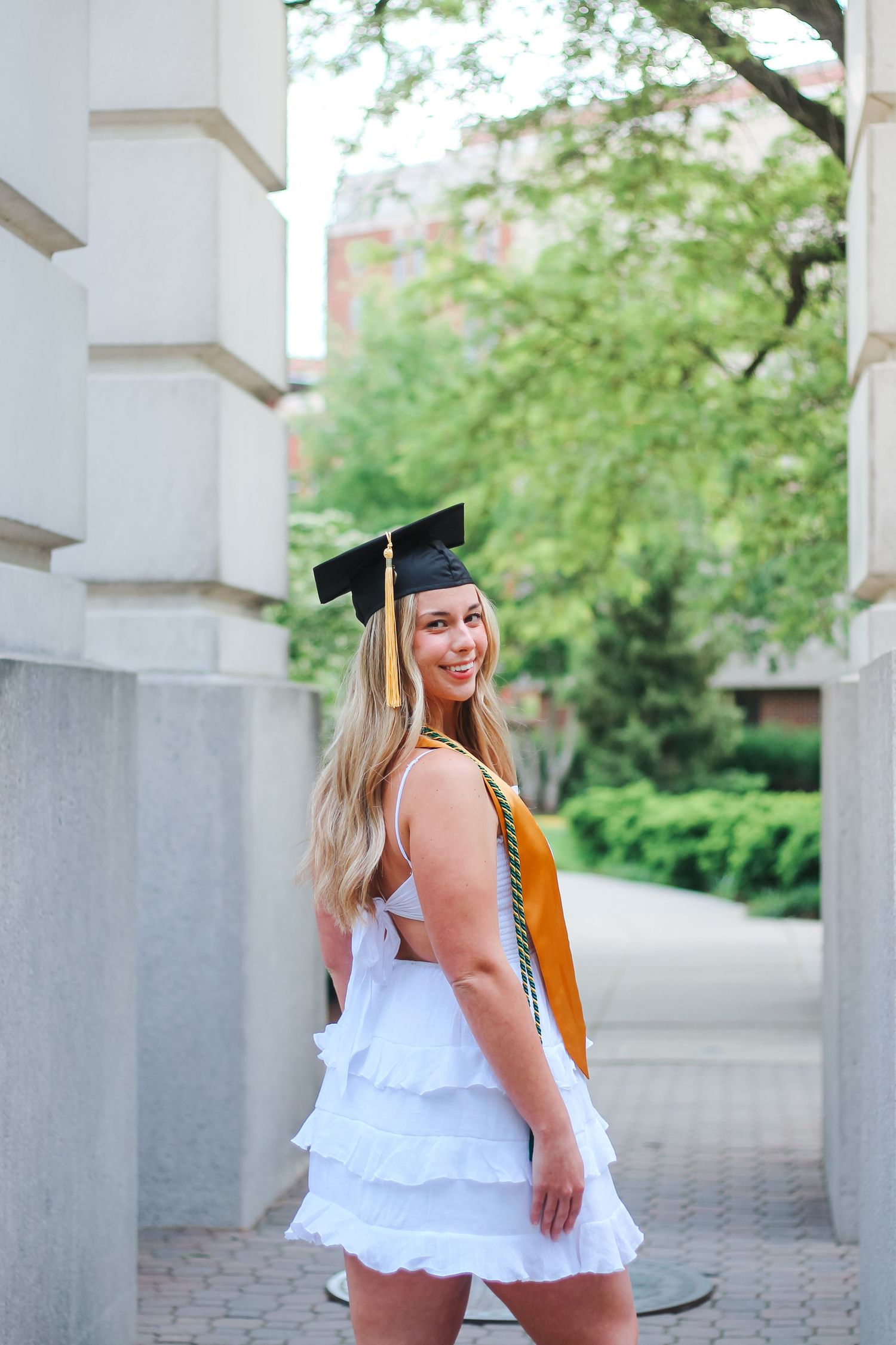 Smiling blonde woman in white dress and black graduation cap with gold stole on campuson Purdue's college campus in West Lafayette