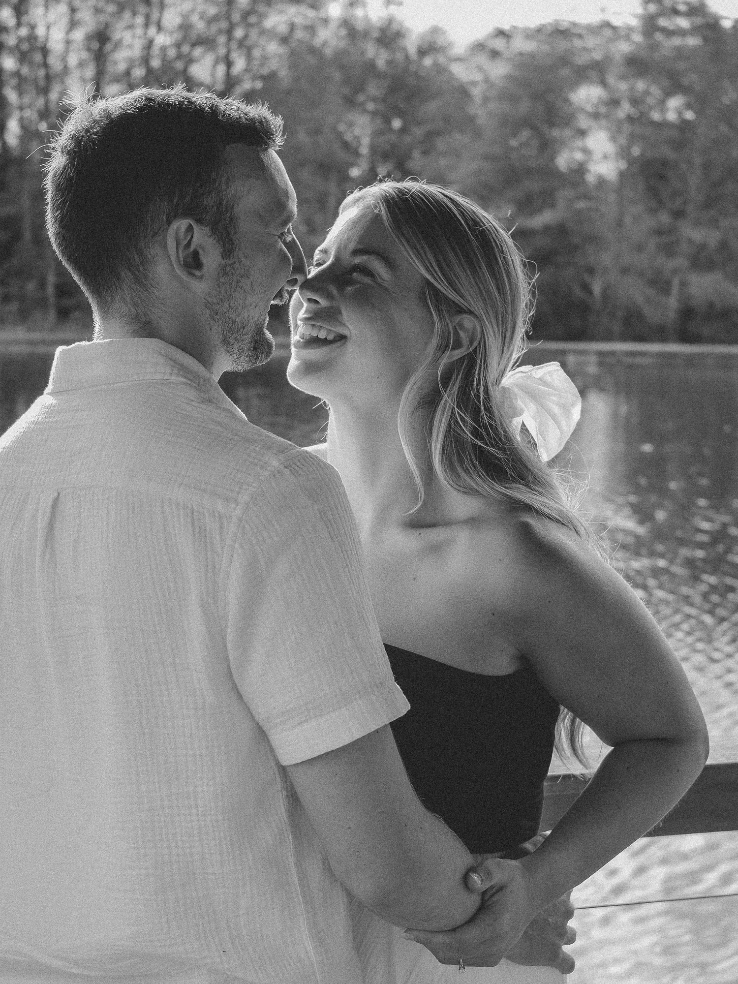 Black and white photo of a laughing woman and man embracing near a serene lake backdrop Hamilton County couples photographer Regan Nichalson
