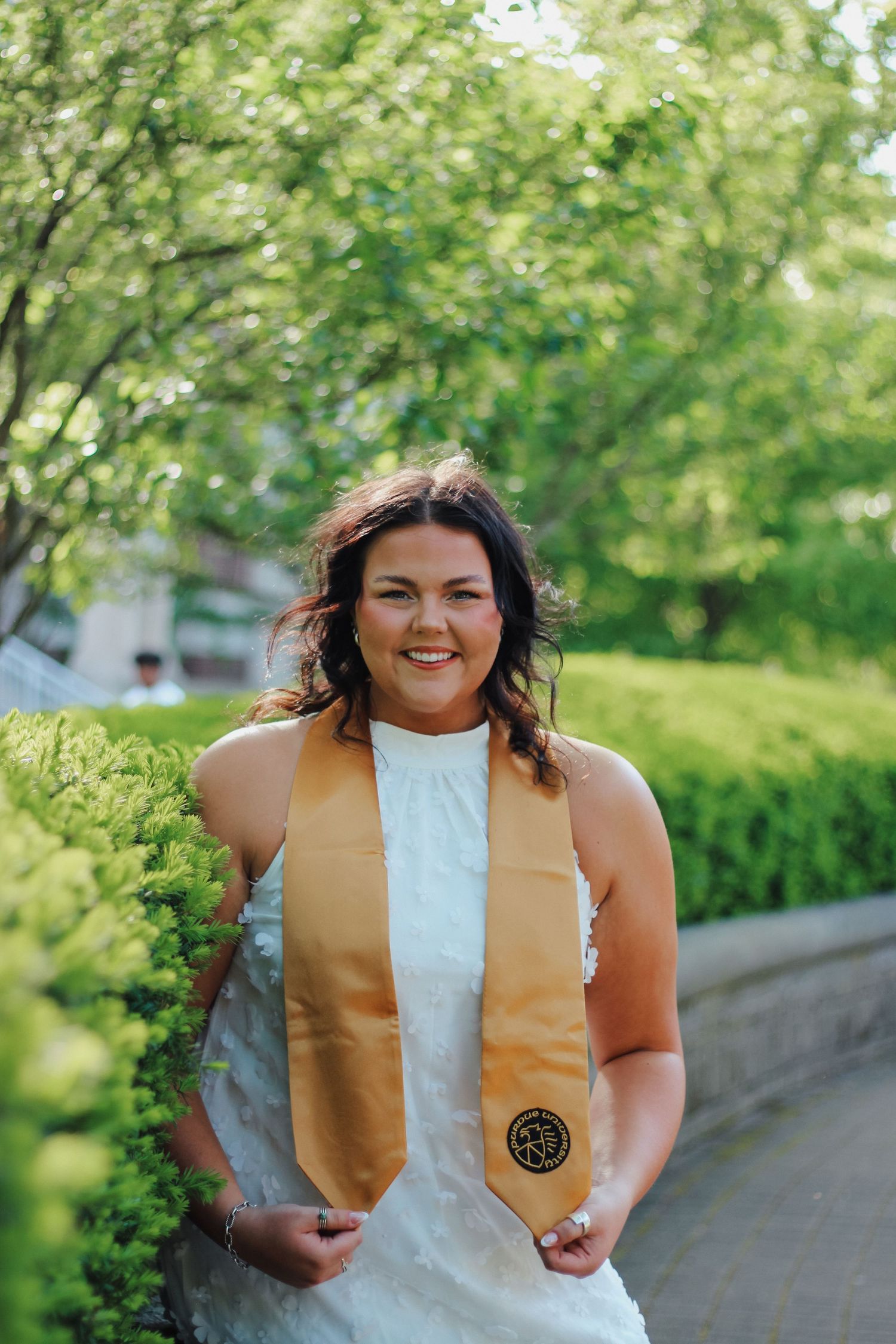 Smiling brunette woman in white dress with gold graduation stole posing along garden path on Purdue's college campus in West Lafayette