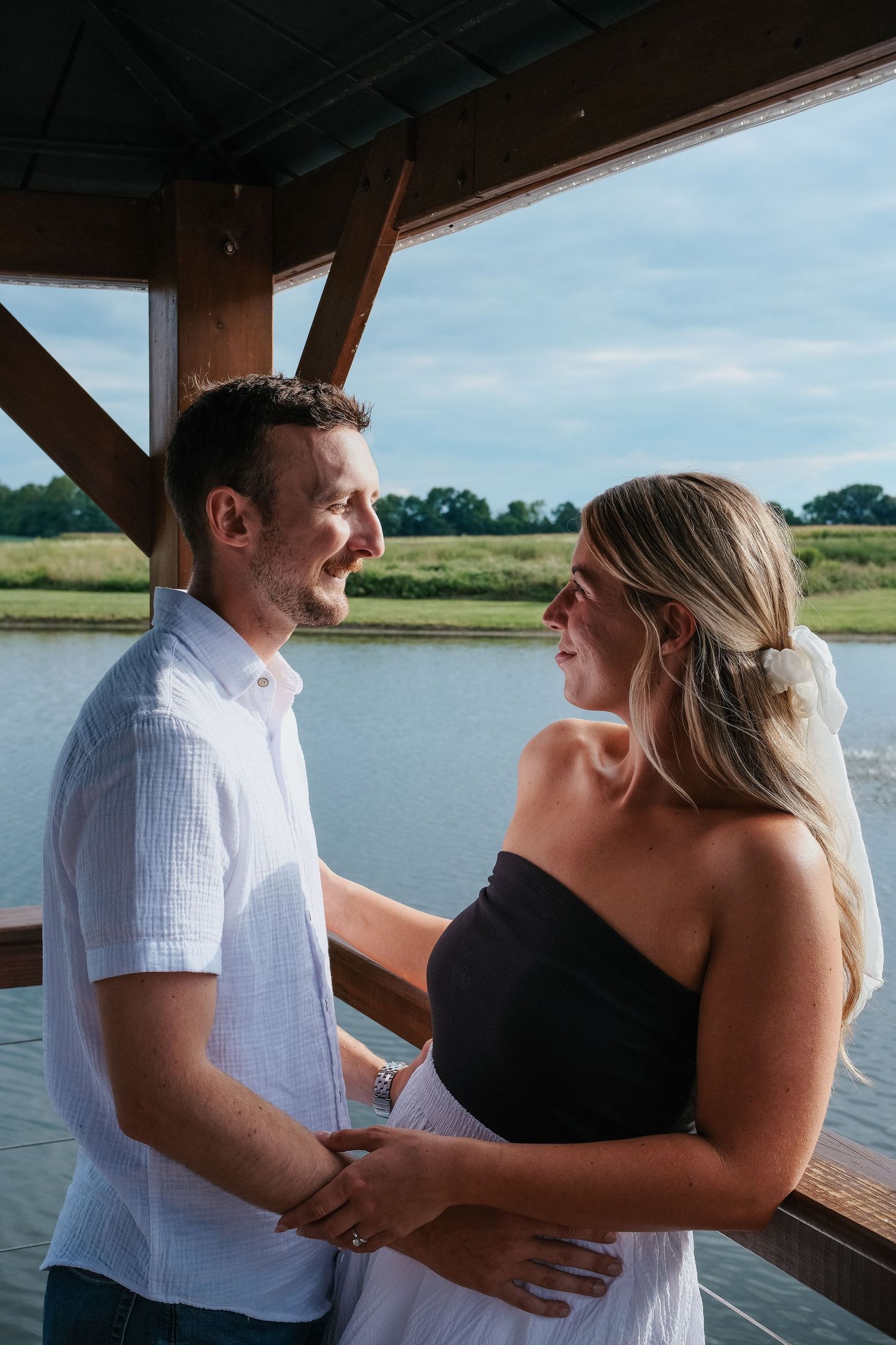 A happy couple holds hands and gazes at each other on a lakeside dock under a wooden gazebo