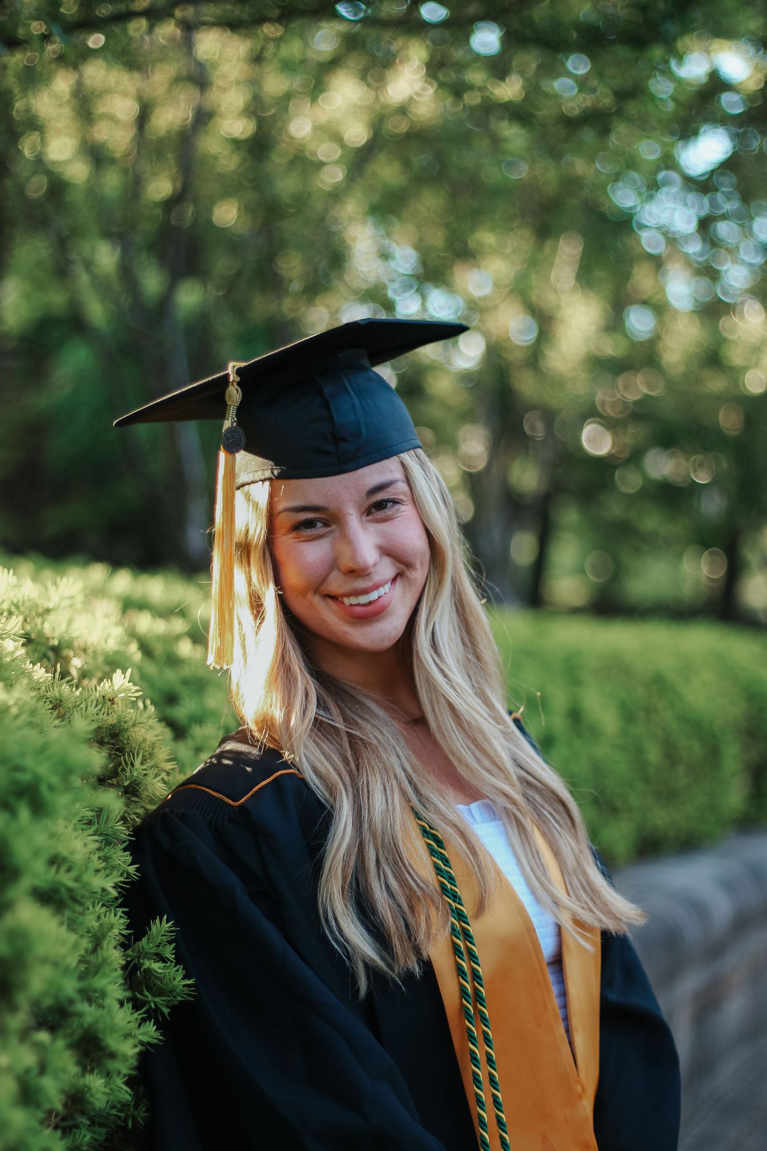 Smiling blonde woman in black graduation cap and gown standing among green bushes outdoors on Purdue's college campus in West Lafayette