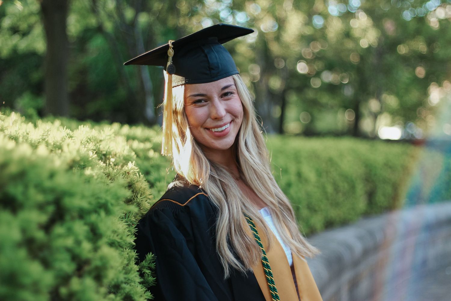 Smiling blonde woman in black graduation cap and gown with gold honor cords outdoors on Purdue's college campus in West Lafayette