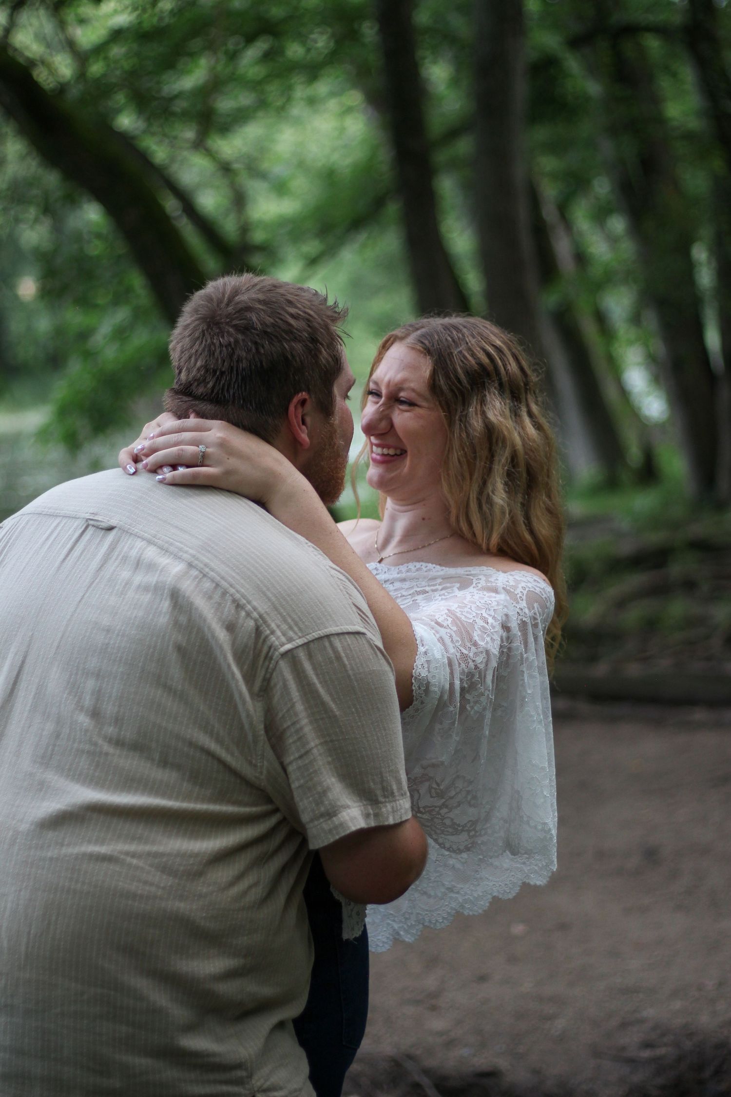 Couple embracing and smiling in a lush green forest setting, woman in white lace top laughing joyfully.