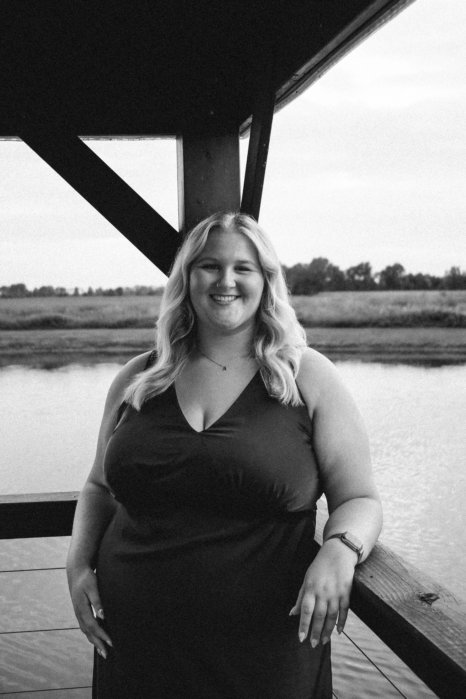 Black and white photo of a cheerful blonde woman in a dark dress posing on a lakeside dock Zionsville family photographer Regan Nichalson