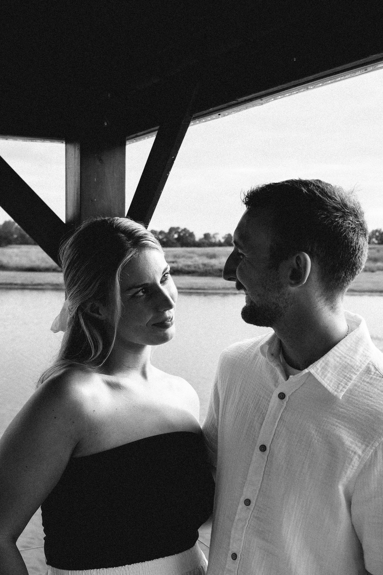 Black and white portrait of a couple gazing at each other under a wooden structure by a lake.